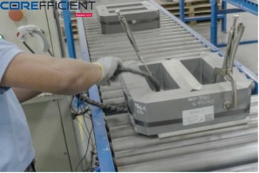 A worker tests dimensional electrical steel coil in manufacturing or assembly. The person is wearing gloves and holding a tool connected to a grey rectangular coil. The conveyor belt aligns similar coils, preparing them for processing or assembly. The logo “COREFFICIENT” is visible at the top left corner.