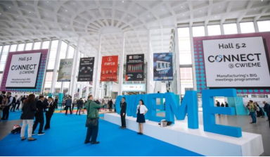 Pictured is a large indoor area with a high ceiling, big windows, and an attractive modern design with white architectural shapes. Big blue letters at the center of the atrium spell “CWIEME.” Lots of people are talking and walking around the space. Banners advertising different companies are hanging from the ceiling.