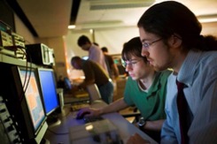 In the foreground, two young men wearing glasses look at connected computer monitors, while in the background other men are at work leaning over a table.