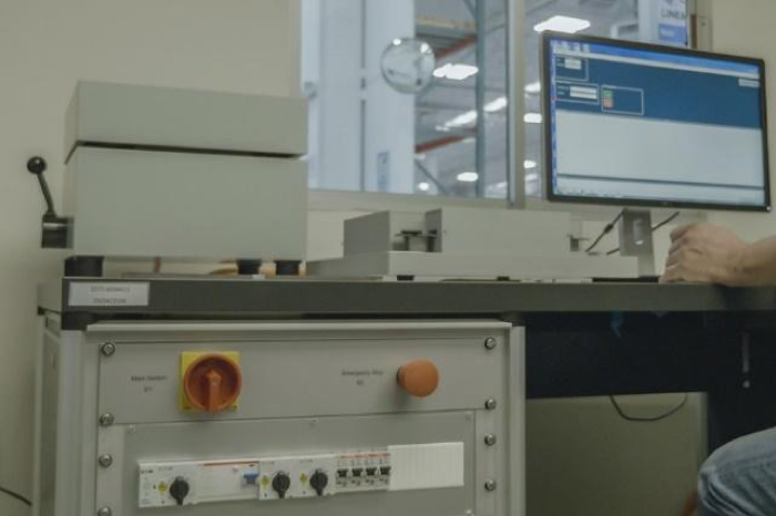 A machine with several knobs and an orange button sits below a table upon which there is plain, off-white machine that resembles a box with a lever and also the hand and knee of a person working a computer behind which is a window that looks towards the interior of a warehouse.
