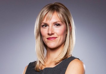 A smiling, brown-eyed Lynn Jurich wearing a charcoal sleeveless dress is standing in front of a gradient grey portrait backdrop her shoulder-length blonde hair beautifully illuminated in the studio lighting.