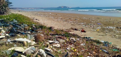 A panoramic picture of a beach littered with plastic and trash as waves crash against the sand.
