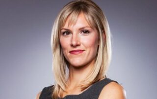 A smiling, brown-eyed Lynn Jurich wearing a charcoal sleeveless dress is standing in front of a gradient grey portrait backdrop her shoulder-length blonde hair beautifully illuminated in the studio lighting.