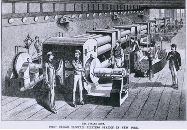 A very old black and white photo of the first Edison Electric lighting station that seems like it is being operated by 6 people in vests. On the bottom of the photo is a text that says, “The Dynamo Room First Edison Electric Lighting Station in New York”.