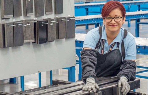 A worker wearing glasses, work gloves, and other protective gear smiles for the camera at a workstation in a factory next to a wall organized with different sizes of metal plates.