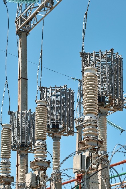This power distribution station has tall ceramic insulators stacked vertically, appearing white and cylindrical. Metal structures are connected to the top of the insulators for electricity transmission, with multiple cables and wires indicating complex circuitry. A metal tower supports the setup against a clear blue sky.
