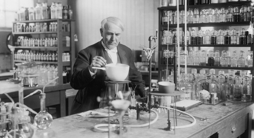 A sepia-toned photograph of Edison tinkering around in his laboratory. Dressed in a fancy, old-fashioned suit, he stands behind a long table with a vintage chemistry set, and behind him are shelves of various chemicals in small glass bottles.
